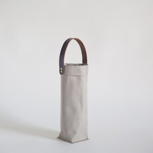 May include: A gray fabric wine bottle carrier with a brown leather handle. The carrier has a rectangular shape and is made of a sturdy material.
