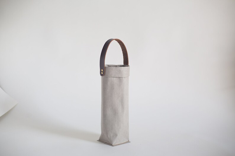 May include: A gray fabric wine bottle carrier with a brown leather handle. The carrier has a rectangular shape and is made of a sturdy material.