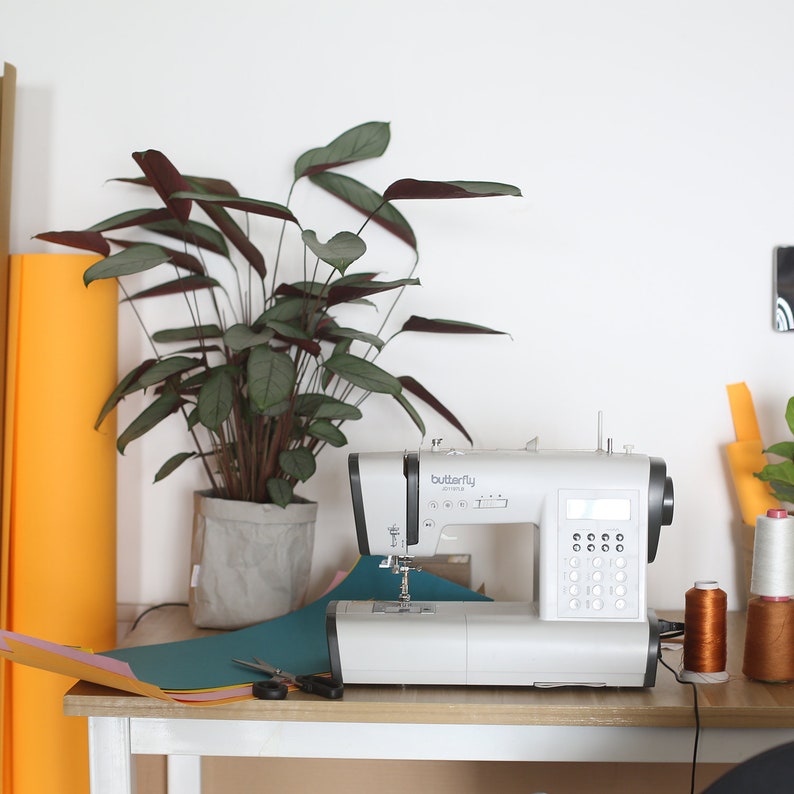 May include: A white sewing machine with the word "butterfly" on it, sitting on a wooden table with a green, blue, and orange piece of fabric, a pair of scissors, and spools of thread. A potted plant with green leaves is in the background.
