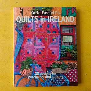 May include: The cover of a book, "Kaffe Fassett's Quilts in Ireland", displays a vibrant quilt design. A bicycle with baskets of vegetables is also shown. The text reads "20 designs for patchwork and quilting".