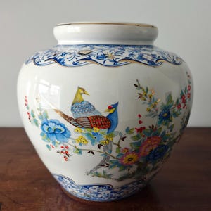 May include: A white ceramic vase featuring a hand-painted design of two colourful pheasants perched among vibrant blue, pink, and yellow flowers and green foliage. The vase has a gold-trimmed rim and decorative blue floral borders.