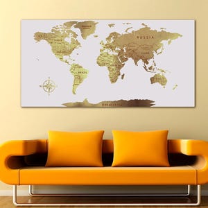 Op de afbeelding: Een goudkleurige wereldkaart op een witte achtergrond, aan de muur bevestigd. De kaart toont landnamen en een windroos. Daaronder is een oranje bank met twee oranje kussens zichtbaar.