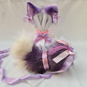 May include: Purple and white furry cat ears and tail with pink and white ribbon accents. The ears are attached to a white headband and the tail has a pink and white ribbon tied around it.
