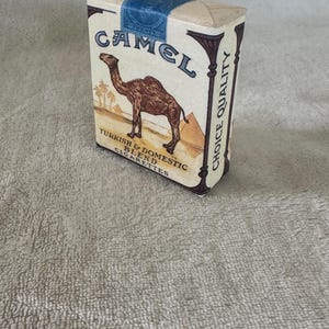 May include: A rectangular Camel cigarette pack with a beige background, featuring a brown camel illustration, pyramids, and palm trees. The text reads Camel, Turkish & Domestic Blend, Choice Quality, and Twenty on a blue tax stamp seal.