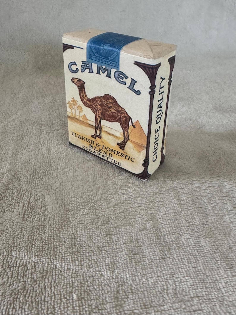 May include: A rectangular Camel cigarette pack with a beige background, featuring a brown camel illustration, pyramids, and palm trees. The text reads Camel, Turkish & Domestic Blend, Choice Quality, and Twenty on a blue tax stamp seal.