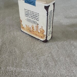 May include: A vintage Camel cigarette pack with a cream-colored paper wrapper, featuring a blue tax stamp on top and a brown illustration of a desert city with domes and minarets. The text reads Don't look for premiums or coupons, as the cost of the tobaccos blended in CAMEL Cigarettes prohibits the use of them.
