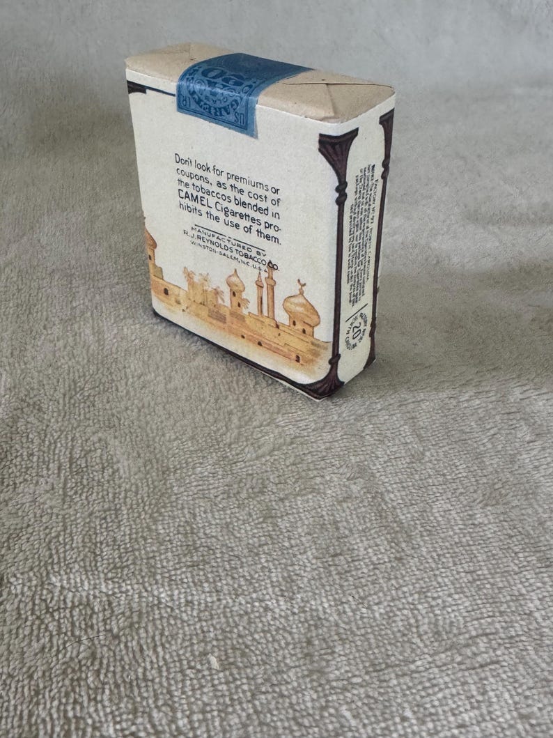 May include: A vintage Camel cigarette pack with a cream-colored paper wrapper, featuring a blue tax stamp on top and a brown illustration of a desert city with domes and minarets. The text reads Don't look for premiums or coupons, as the cost of the tobaccos blended in CAMEL Cigarettes prohibits the use of them.
