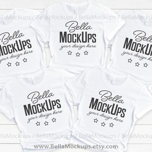 Pode incluir: Cinco camisetas brancas com o texto "Bella Mockups" e "your design here" impresso nelas. As camisetas estão empilhadas em uma superfície de madeira branca.