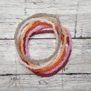 May include: A set of six colorful beaded bracelets in shades of gray, white, peach, orange, pink, and burgundy.