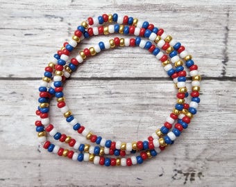 May include: A beaded waist chain featuring red, white, blue, and gold beads. The beads are strung together to form a long, flexible strand. The waist chain is designed to be worn around the waist as a decorative accessory.