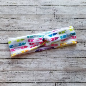 May include: A white headband with a colorful crayon pattern. The headband is twisted and has a knot in the center.
