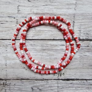 May include: A multi-strand beaded necklace with red, pink, and white beads. The necklace is made of small, round beads and is arranged in a striped pattern.