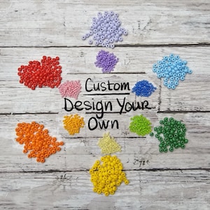 May include: An assortment of colorful beads in red, orange, yellow, green, blue, and purple arranged on a wooden surface. The text "Custom Design Your Own" is in the center, suggesting a DIY craft project or jewelry making supplies.
