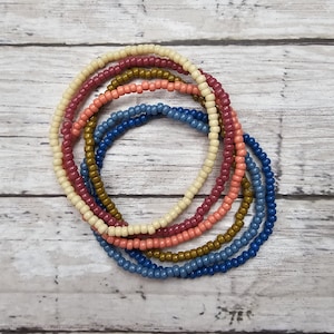 May include: A set of six beaded bracelets in shades of cream, peach, rust, gold, and blue. The bracelets are stacked on top of each other, creating a colorful and layered look.
