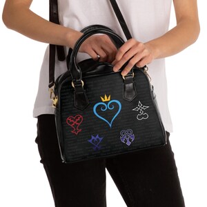 Puede incluir: Bolso de mano negro con asa superior y bandolera. El bolso presenta varios diseños gráficos coloridos, incluido un corazón azul con una corona y otros símbolos. El fondo tiene texto blanco pequeño.