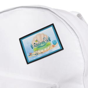 May include: White backpack with a rectangular patch featuring an ocean-themed design. The patch has a light blue background with a seashell, seaweed, and the word "Atlantica". A trident and coral are also visible.