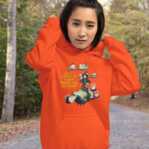 May include: Vibrant orange hoodie featuring a witch-themed graphic with the text "Now where did I put that eye of newt?" The design includes a witch, cauldron, and potion ingredients. The hoodie is made of a soft material.