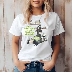 May include: White t-shirt with a whimsical witch-themed graphic. The design features a witch in a black dress and hat, a bubbling cauldron, and the text "Now... where did I put that eye of newt?"