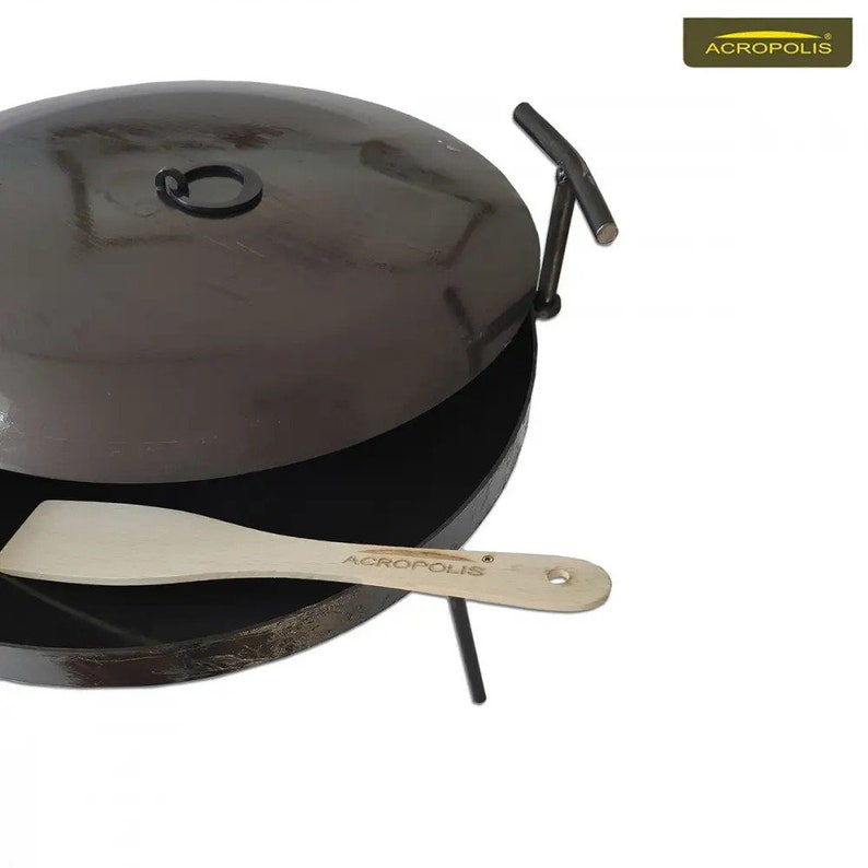 May include: A black metal pan with a lid and a wooden spatula. The pan has a handle on the lid and a stand to hold it up. The spatula has the word "ACROPOLIS" printed on it.