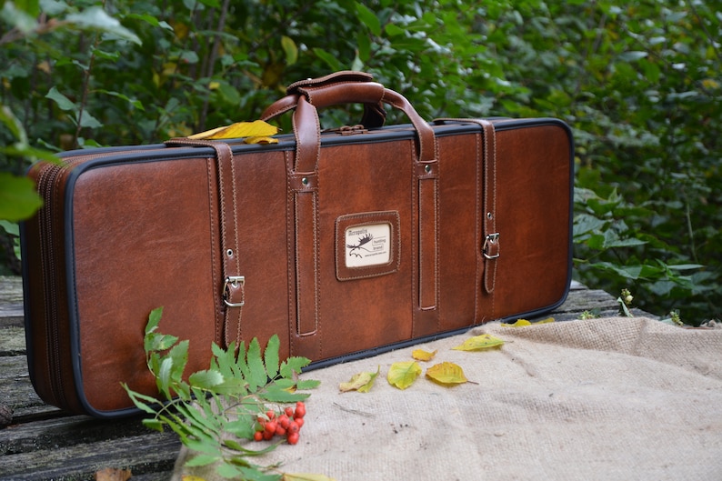 May include: A brown leather gun case with a carrying handle and two buckles. The case has a label with the text "Borealis Hunting Outfitters" on it.