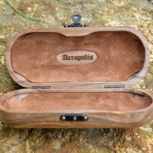 May include: A wooden box with a brown suede interior. The box has a hinged lid and a metal clasp. The box is engraved with the word "Acropolis".