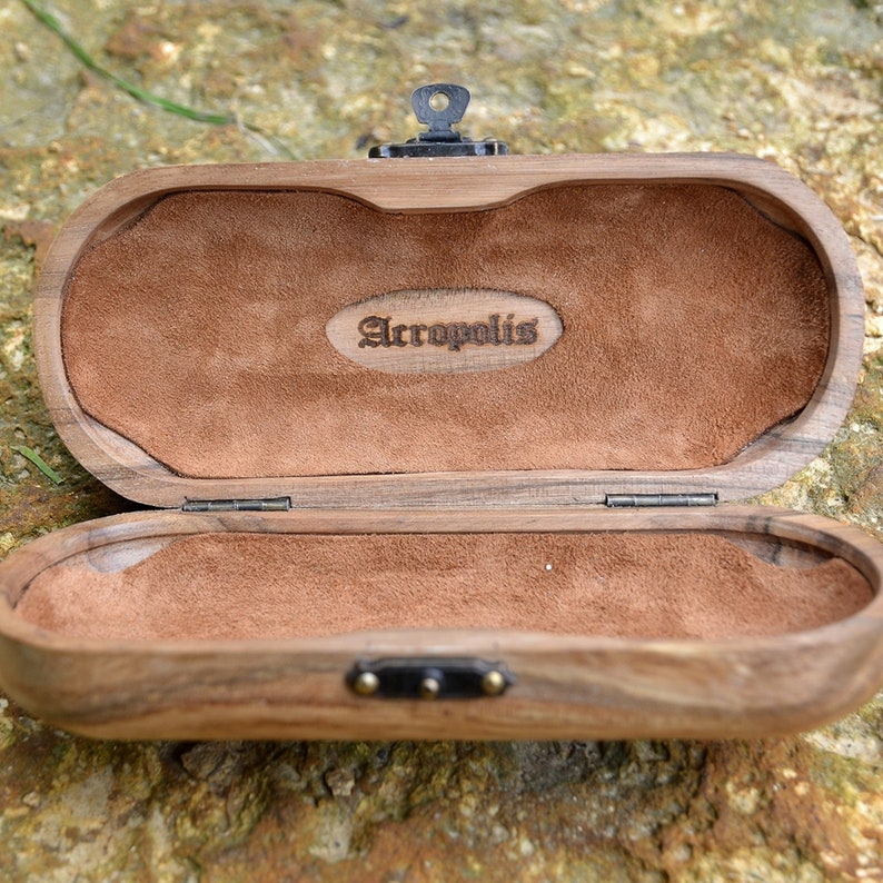 May include: A wooden box with a brown suede interior. The box has a hinged lid and a metal clasp. The box is engraved with the word "Acropolis".