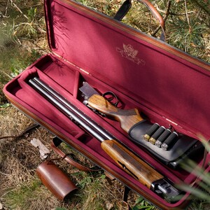 May include: A brown leather gun case with a red interior. The case is open and a shotgun is inside. The shotgun has a wooden stock and a black metal barrel. There are also several shotgun shells in the case.
