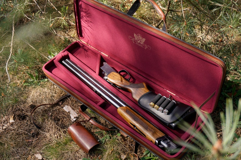 May include: A brown leather gun case with a red interior. The case is open and a shotgun is inside. The shotgun has a wooden stock and a black metal barrel. There are also several shotgun shells in the case.
