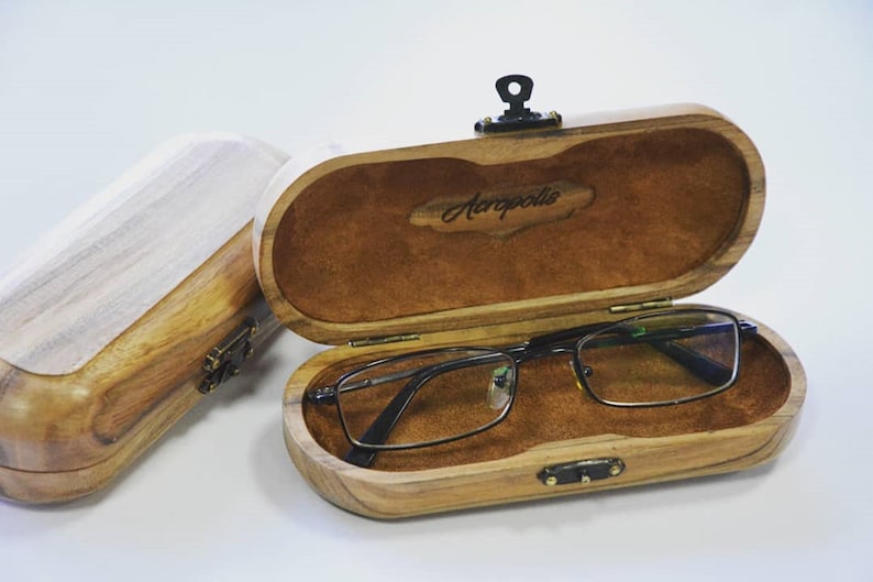 May include: Two wooden eyeglass cases, one open and one closed. The open case has a brown suede interior and a pair of eyeglasses inside. The case has a brass latch and the text "Aeropolis" is engraved on the lid.