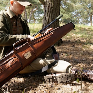 May include: A brown leather rifle case with a zipper closure and a shoulder strap. The case is open, revealing a rifle. A person is sitting on the ground in a forest, wearing a green jacket and a brown cap. A pair of brown leather boots are also visible.