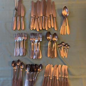 May include: A large collection of silver-toned stainless steel flatware including dinner knives, forks, soup spoons, teaspoons, and a serving fork. The set features a simple, smooth handle design with rounded ends, arranged in organized groups.