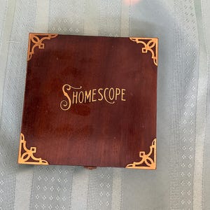 May include: A square, wooden box with a dark brown finish and gold-colored corner accents. The word "HOMESCOPE" is written in gold script on the top. The box is closed and sits on a light blue patterned surface.