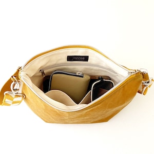May include: A mustard yellow canvas shoulder bag with a white and black checkered strap. The bag is open and has multiple pockets inside. The bag is lined with a cream colored fabric.