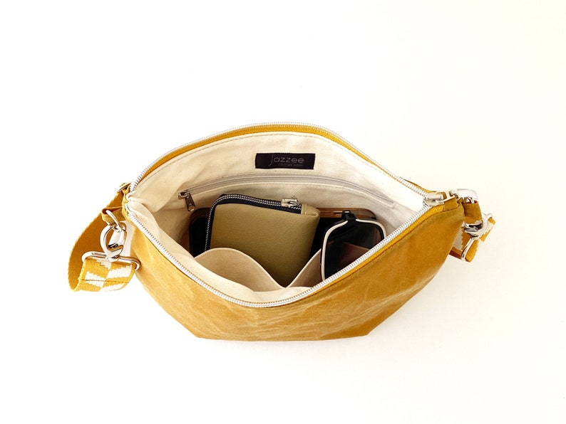 May include: A mustard yellow canvas shoulder bag with a white and black checkered strap. The bag is open and has multiple pockets inside. The bag is lined with a cream colored fabric.