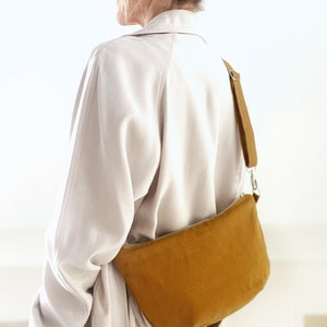 May include: A mustard yellow suede crossbody bag with a long adjustable strap. The bag is worn by a person wearing a light beige jacket.