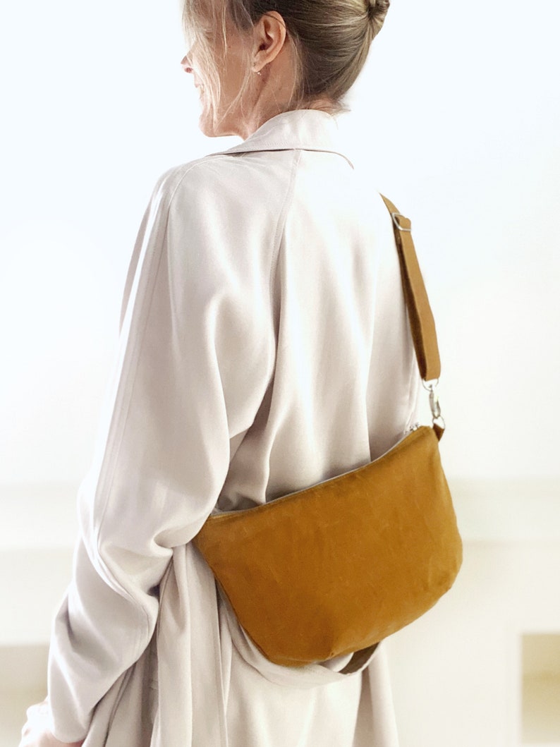 May include: A mustard yellow suede crossbody bag with a long adjustable strap. The bag is worn by a person wearing a light beige jacket.