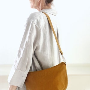 May include: A brown canvas shoulder bag with a long strap. The bag is worn by a person wearing a white linen shirt.