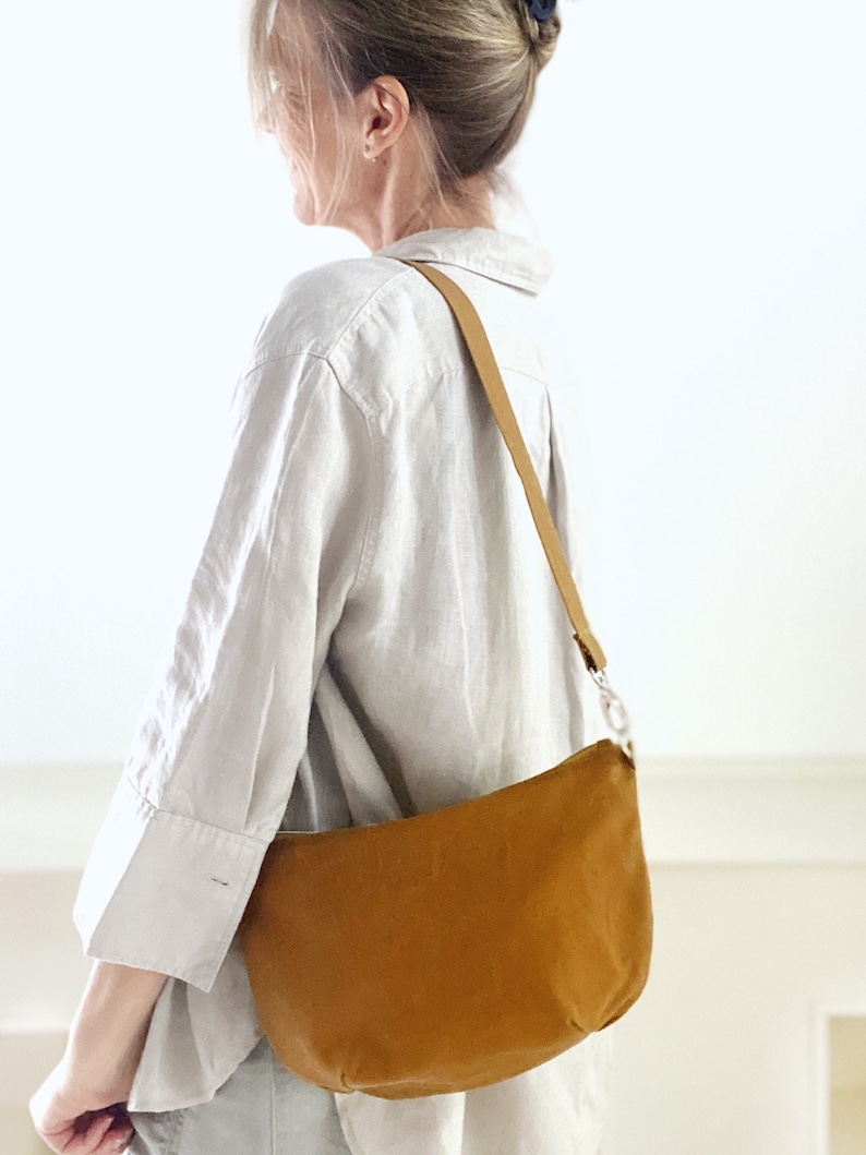 May include: A brown canvas shoulder bag with a long strap. The bag is worn by a person wearing a white linen shirt.