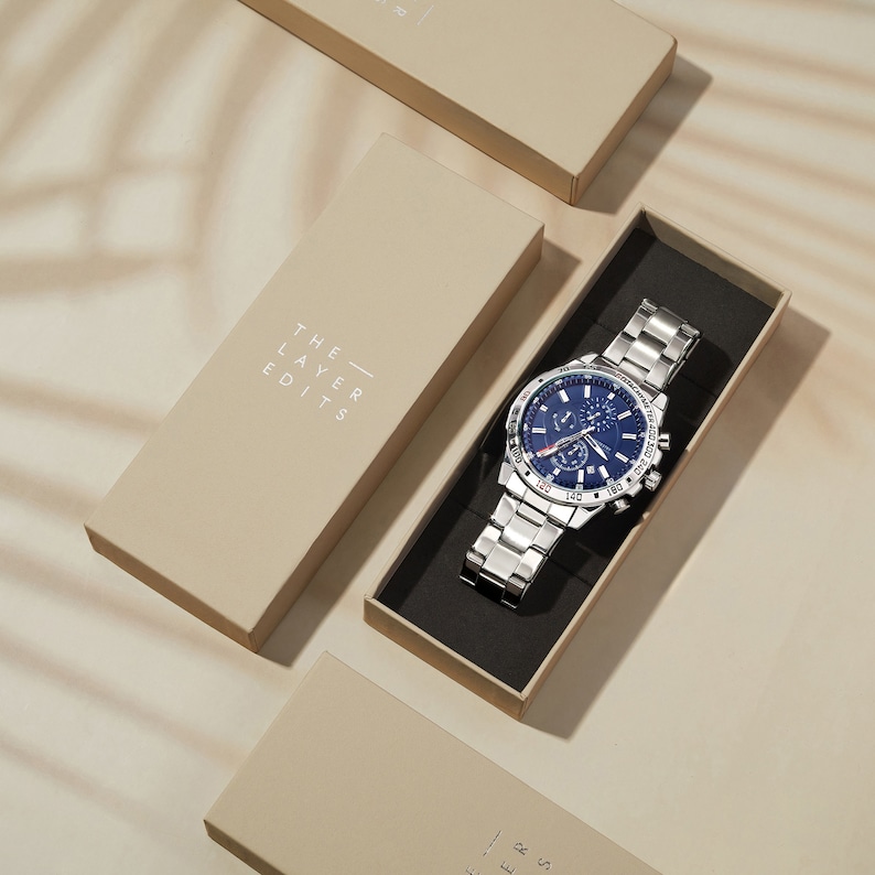 May include: A silver watch with a blue face and silver hands. The watch has a chronograph function and a date window. The watch is in a box with the words "The Layer Edits" printed on the side.