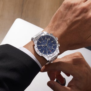 May include: A silver chronograph wristwatch with a blue face and red accents. The watch has a stainless steel band and a tachymeter scale around the outer edge of the face.