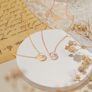 Puede incluir: Dos delicados collares con colgantes. Uno es dorado con un colgante redondo, el otro es de oro rosa con un colgante cuadrado. Ambos colgantes presentan una piedra central transparente rodeada de piedras m&aacute;s peque&ntilde;as.