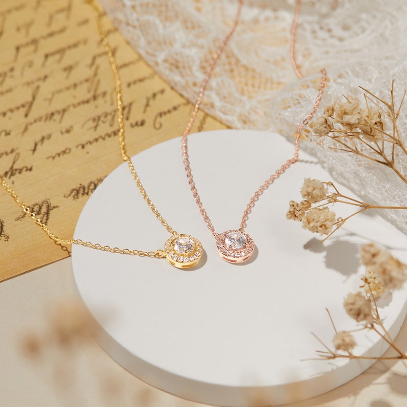 Puede incluir: Dos delicados collares con colgantes. Uno es dorado con un colgante redondo, el otro es de oro rosa con un colgante cuadrado. Ambos colgantes presentan una piedra central transparente rodeada de piedras m&aacute;s peque&ntilde;as.