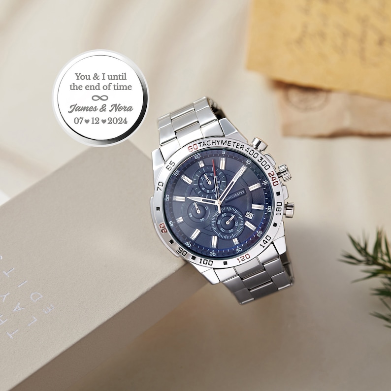 May include: A silver chronograph watch with a blue face and a silver band. The watch has a tachymeter scale around the edge of the face. The watch is engraved with the text "You & I until the end of time" and "James & Nora 07 12 2024".