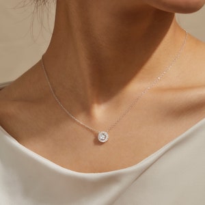Puede incluir: Un delicado collar de plata con un colgante redondo con una piedra transparente central rodeada de piedras m&aacute;s peque&ntilde;as. El colgante cuelga de una fina cadena, creando un aspecto cl&aacute;sico y elegante. El collar se muestra sobre un fondo neutro.