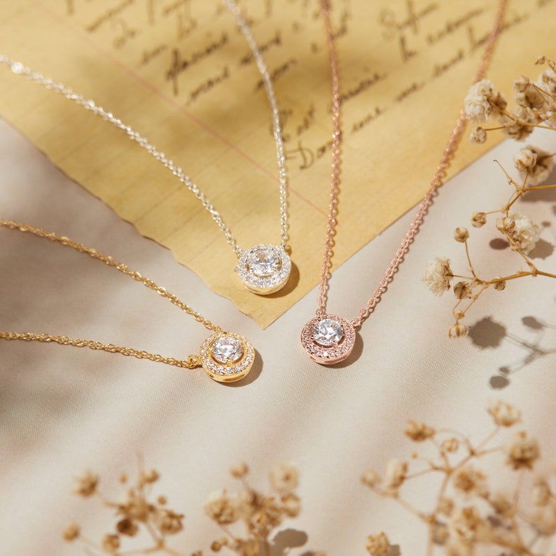 Puede incluir: Cuatro collares con una piedra central clara y redonda rodeada de piedras m&aacute;s peque&ntilde;as. Los collares son dorados, plateados y oro rosa. Las cadenas son delicadas, sobre un fondo de papel vintage.
