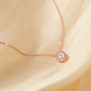 Puede incluir: Un collar de oro rosa con un colgante redondo de piedra preciosa transparente. El colgante est&aacute; rodeado de piedras preciosas transparentes m&aacute;s peque&ntilde;as. La delicada cadena descansa sobre una tela color crema similar al sat&eacute;n.