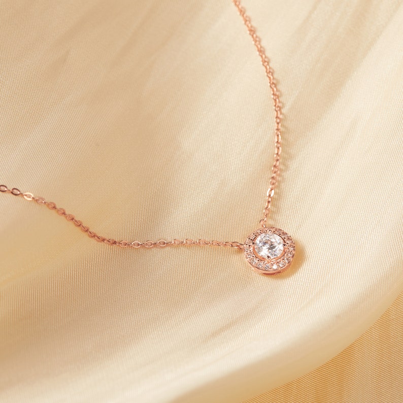 Puede incluir: Un collar de oro rosa con un colgante redondo de piedra preciosa transparente. El colgante est&aacute; rodeado de piedras preciosas transparentes m&aacute;s peque&ntilde;as. La delicada cadena descansa sobre una tela color crema similar al sat&eacute;n.