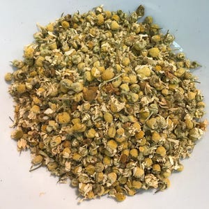 May include: A pile of dried chamomile flowers consisting of small, round, yellow centres surrounded by delicate, pale white petals. The dried botanical material features a mix of whole flower heads and fragmented floral parts in shades of gold, cream, and light green, creating a textured, organic appearance.