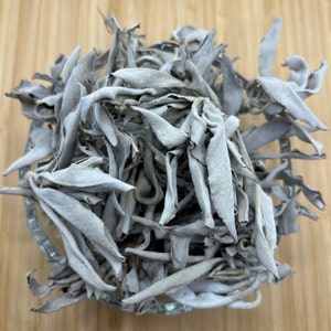 May include: A glass bowl containing dried white sage leaves. The leaves are a pale grey colour, with a textured surface and curled edges. The bowl rests on a light brown wooden surface.