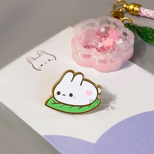 May include: A white bunny rabbit with pink ears and a pink heart on its chest is sitting on a green leaf. The bunny is a gold-colored enamel pin.